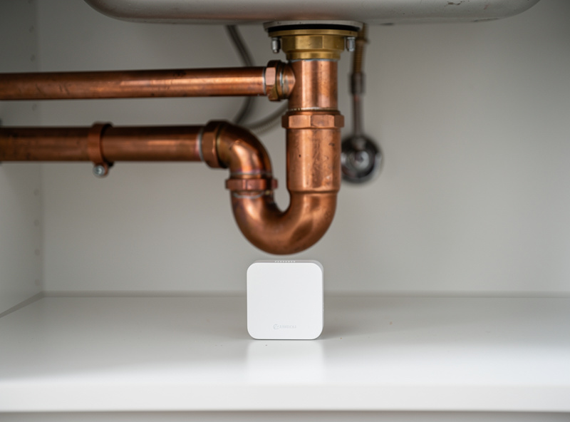 Smart water leak detector sensor installed under kitchen sink with visible plumbing pipes