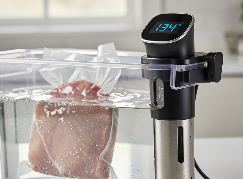 Smart sous vide circulator clipped to clear container with vacuum-sealed steak inside, smartphone showing app