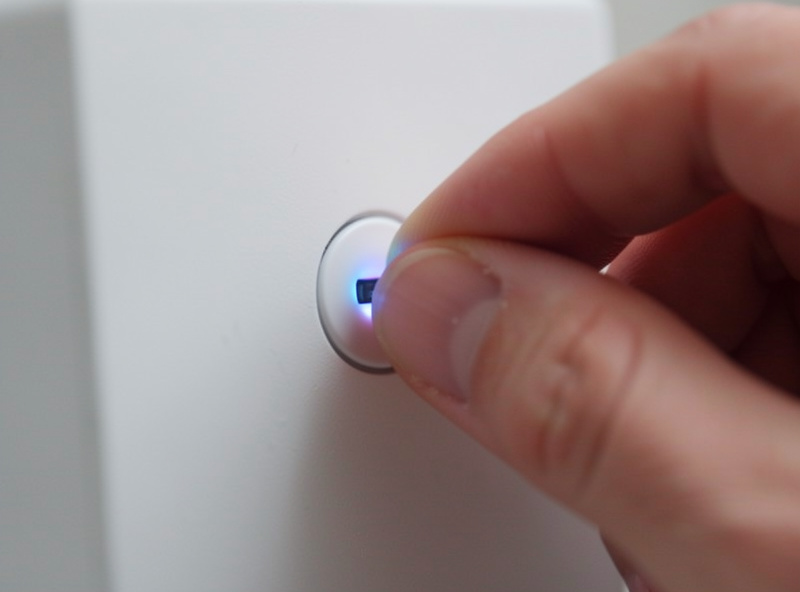 A finger holding down the reset button on a smart home device until its indicator light blinks