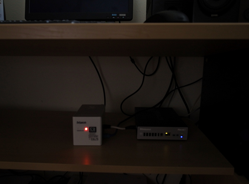 Small consumer UPS battery backup wired to a mini-PC and network switch under a desk with a glowing status LED
