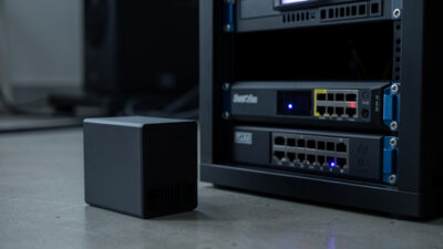 Small consumer UPS battery backup beside a network rack with a mini-PC and switch, status LED glowing in a dim room