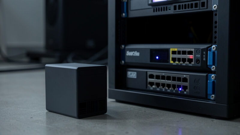 Small consumer UPS battery backup beside a network rack with a mini-PC and switch, status LED glowing in a dim room