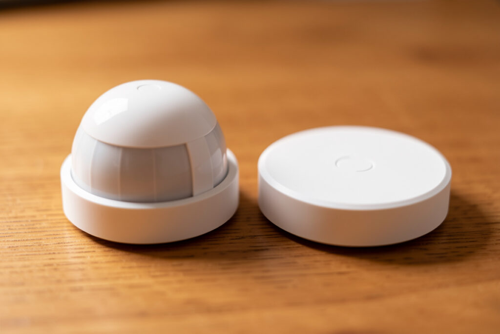 Two smart-home sensors side by side, a dome PIR motion sensor and a flat mmWave radar sensor on wood