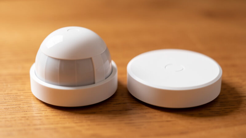 Two smart-home sensors side by side, a dome PIR motion sensor and a flat mmWave radar sensor on wood
